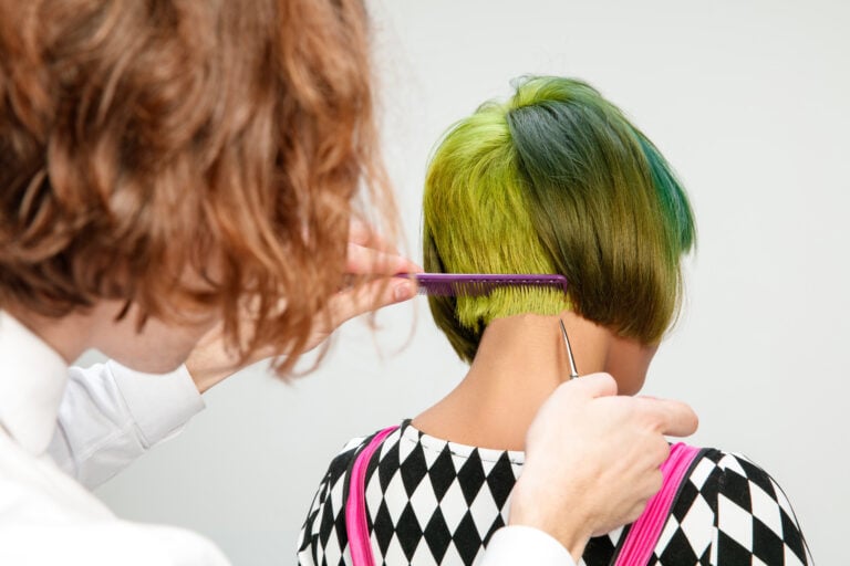 Picture showing adult woman at the hair salon. Studio shot of graceful young girl with stylish short haircut and colorful hair on gray background and hands of hairdresser.